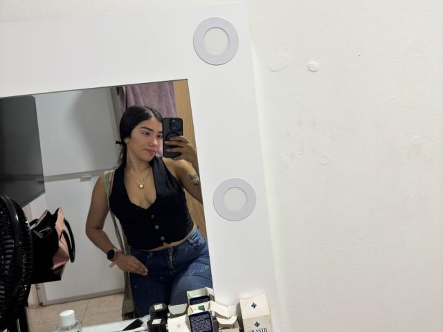 SharonKlart with small tits, wearing a black halter top and blue jeans, taking a mirror selfie, showing a pouty face, with a pink bag and makeup on the counter.