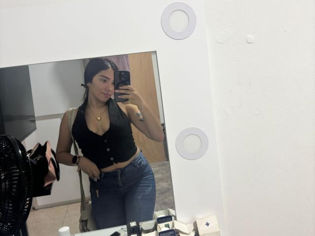 SharonKlart taking a mirror selfie, wearing a black halter top showing her small tits, blue jeans, and a gold necklace. Her long black hair is down, with a tattoo on her arm.