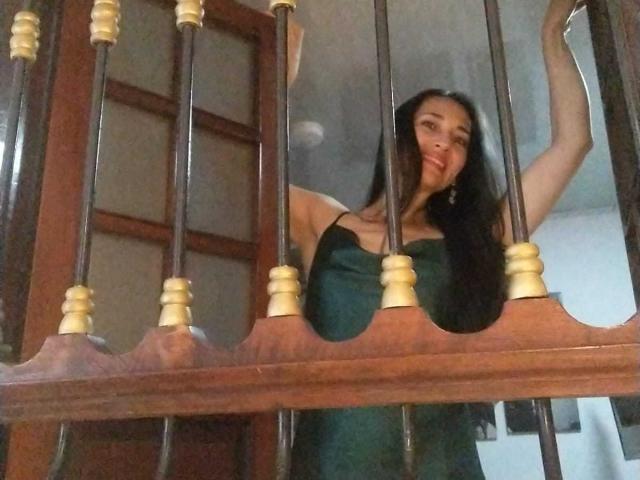 MiaMott, con i suoi lunghi capelli scuri, il vestito verde e gli orecchini, si sporge attraverso delle sbarre di legno e metallo, sorridendo, con le braccia alzate, mostrando leggermente il seno..