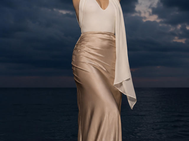 NeldaBeierle in a white halter dress with a flowing scarf and a shiny, beige satin skirt, standing by dark water, showing her ass and a hint of tits.