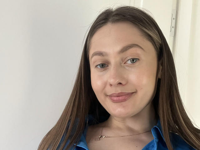 MisVasilisa ha lunghi capelli castani, occhi verdi e un leggero sorriso, indossa una camicia blu con una sottile collana, mostrando una pelle liscia e chiara e un viso carino..