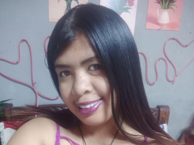 MariahSquirting69 con capelli neri lucidi, rossetto viola, che sorride, mostrando i denti, indossa un reggiseno rosa, una collana nera, uno sfondo con un muro grigio, un cordoncino rosso a forma di cuore e decorazioni murali floreali..