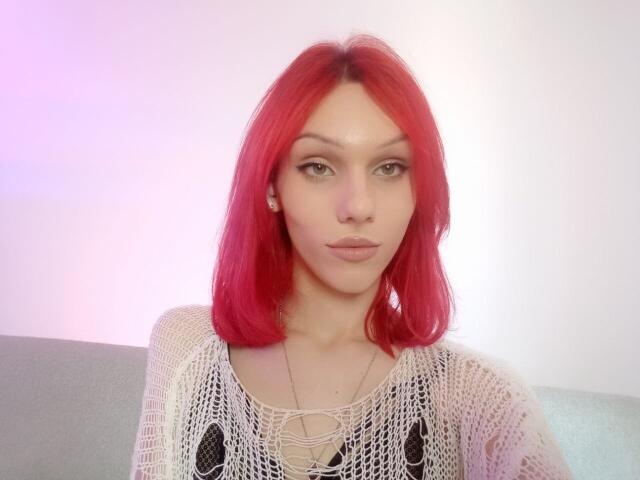 KiranaHayes with red hair, wearing a white knit top, has small tits, and a neutral expression, sitting on a gray couch, looking at the camera.