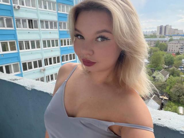 ChuHeal, con i capelli biondi, gli occhi azzurri e il rossetto rosso, indossa una maglietta grigia a spalle scoperte, che mette in mostra il suo seno. È in piedi su un balcone con un edificio blu sullo sfondo..