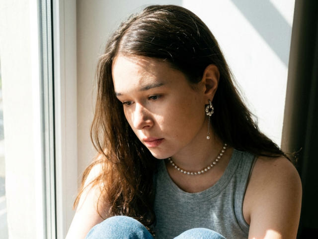 Jannet Rundahl, met lang bruin haar, een grijze tanktop, een parelketting en oorbellen, kijkt naar beneden met een nadenkende uitdrukking, zittend bij een raam, en draagt blauwe jeans..