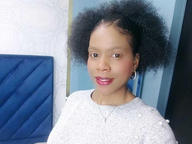 SarahiPusey with big black afro, brown eyes, pink lipstick, gray textured top, silver hoop earrings, and a blue quilted chair in the background.