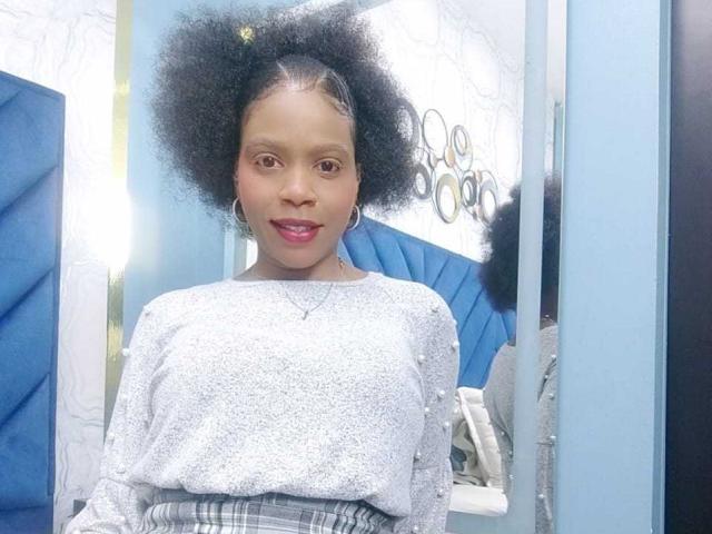 SarahiPusey with medium brown skin, curly black hair, small nose, pink lipstick, wearing a silver sparkly top and plaid skirt, standing in front of a mirror.