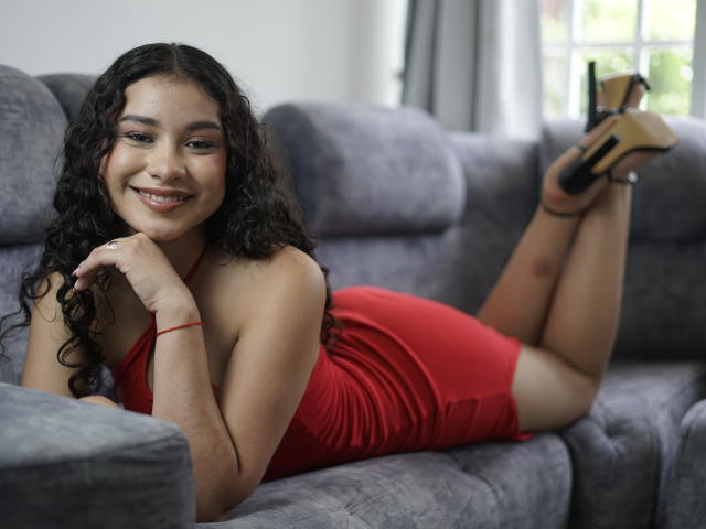 HemMastar, avec ses cheveux noirs bouclés, est allongée sur un canapé gris, vêtue d'une robe rouge, souriante, les jambes en l'air, montrant ses fesses, chaussée de talons beiges, et portant un bracelet rouge à son poignet droit..