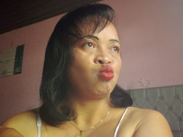 Rahel69 mit geschwollenen, braunen Augen, schwarzem, schulterlangem Haar, einem weißen Top mit Spaghetti-Trägern und einer goldenen Halskette, die ihren Hintern für einen Kuss in einem rosa gefliesten Raum präsentiert..