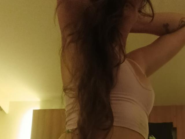 NightmareRaven con cabello castaño largo y ondulado, vistiendo un top deportivo rosa, levantando los brazos, mostrando el perfil lateral, con un tatuaje visible en el brazo derecho. Iluminación tenue y amarilla..