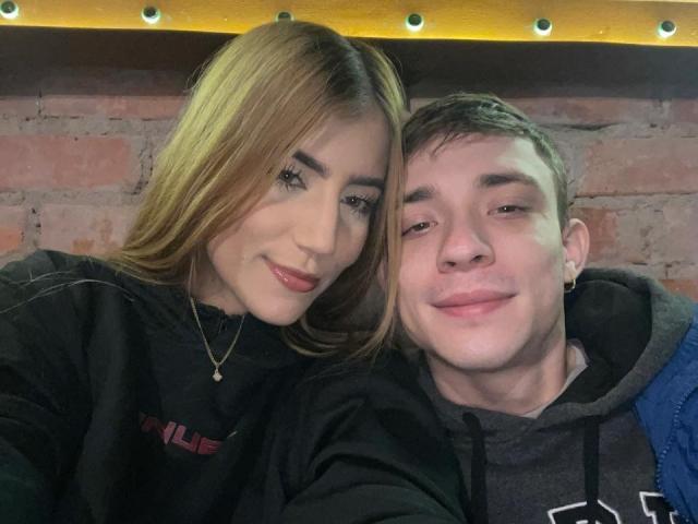 SecretLoversVIP with blonde hair, red lipstick, and black hoodie, next to SecretLoversVIP with short brown hair, ear piercing, and gray hoodie, smiling closely.