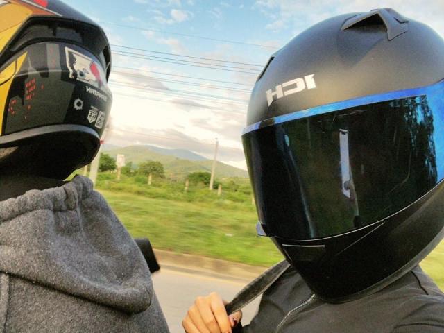 SecretLoversVIP in black HD1 helmet, black jacket, right hand on chest, next to SecretLoversVIP in gray hoodie, yellow and black helmet, road blurred in background.