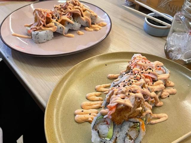 Two plates of sushi with creamy orange sauce, black sesame seeds, and chopped green onions on beige and pink plates, showing different amounts of sauce.
