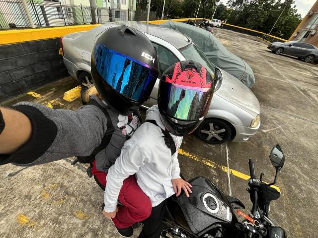 SecretLoversVIP with shiny blue and pink visor helmets, white shirt, red pants, and black motorcycle, standing next to covered silver car, gray-wearing arm in shot, concrete parking lot.