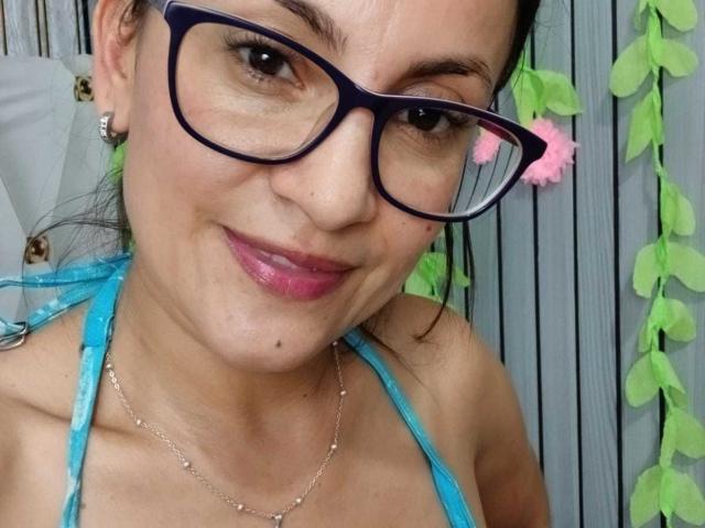 SolaraDubois with glasses, pink lipstick, and blue halter top, showing off her tits and a small beauty mark on her chin, smiling at the camera.