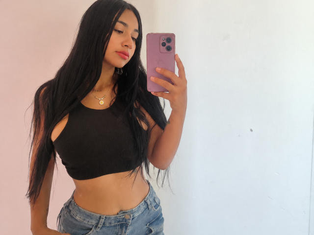 SexyCandyBabe69 mit langen, schwarzen Haaren, trägt ein schwarzes Crop-Top und blaue Jeans, hält ein rosa Handy, zeigt ihren flachen Bauch und ihre kleinen Brüste und steht in einem weißen Raum..
