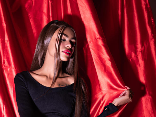 CrystalDavids, met lang bruin haar, een zwarte top en rode lippenstift, houdt een rode satijnachtige gordijn vast en toont een tatoeage op haar borst. Ze is aantrekkelijk en kijkt naar beneden met een lichte glimlach..