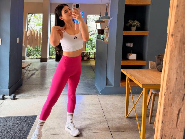 Paulina, con lunghi capelli scuri, un reggiseno sportivo bianco, leggings rosa a vita alta, scarpe da ginnastica bianche e calze a righe, sta scattando un selfie allo specchio, mostrando il suo addome e i glutei scolpiti..