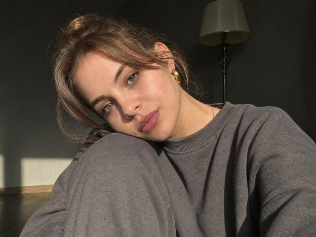 Lilia Pochatko, mit dem Kopf auf ihrem Knie, trägt einen grauen Pullover, goldene Ohrringe und hat ein leichtes Lächeln. Ihr braunes Haar ist zu einem unordentlichen Dutt gebunden, und das Sonnenlicht scheint auf ihr Gesicht..