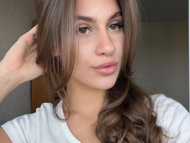 EllenKienbaum has brown eyes, full pink lips, and wavy brown hair, wearing a white shirt, with her hand in her hair, looking directly at the camera.