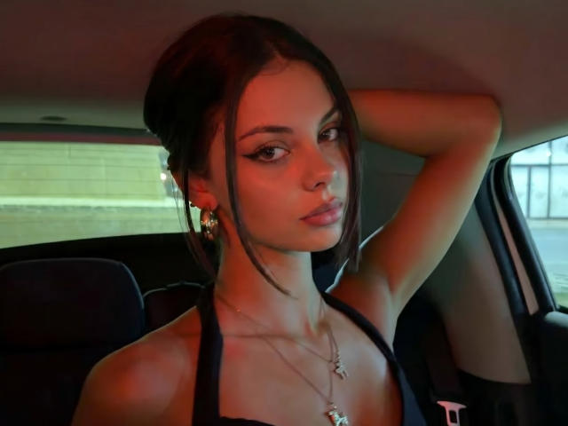 Lilia Pochatko, con i capelli scuri, con una mano alzata, indossa un top nero con spalline che mette in mostra il seno, orecchini dorati, una collana e guarda di lato, seduta in un'auto..