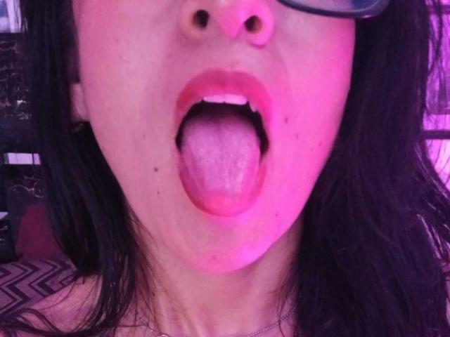 Le visage de ZarayFeet, illuminé d'une teinte rosée, avec ses cheveux noirs, ses lunettes, et sa langue complètement dehors, montrant sa langue rose et ses petites dents blanches, la bouche légèrement ouverte, révélant un peu de ses dents du haut..