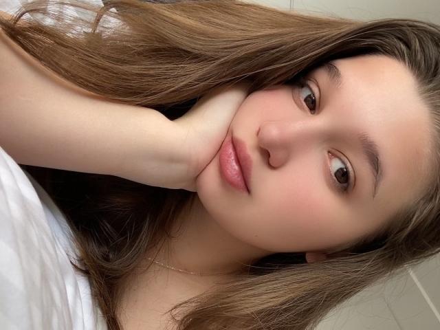 MissKuromi with long brown hair, brown eyes, and pink lips, resting her cheek on her hand, wearing a white shirt, showing a cute, relaxed expression.