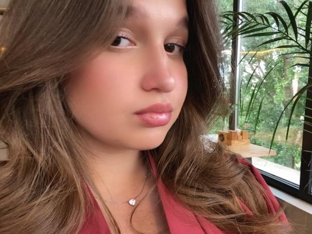 MissKuromi's face, with brown wavy hair, pink lips, and a small silver necklace, showing a slight pout, wearing a red top, in a bright, green-filled room.