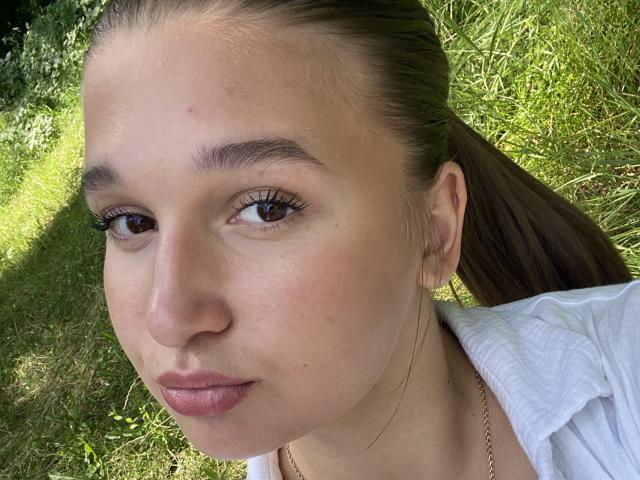 MissKuromi's face, showing her brown eyes, pinkish lips, and slight pimple on her left cheek. She has light skin, brown hair in a ponytail, wearing a white shirt.