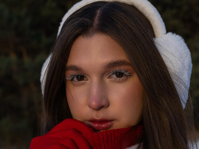 MissKuromi has brown eyes, long brown hair, and is wearing a white fuzzy hat and red sweater, with her hand covering her lower face. Her lips are slightly parted.