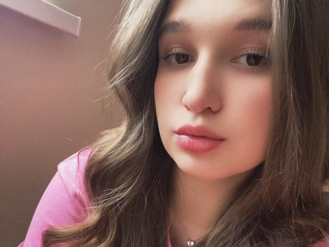 MissKuromi has wavy brown hair, pink lips, and a cute nose with a small nose ring. She's wearing a pink shirt and has a necklace with a small charm.