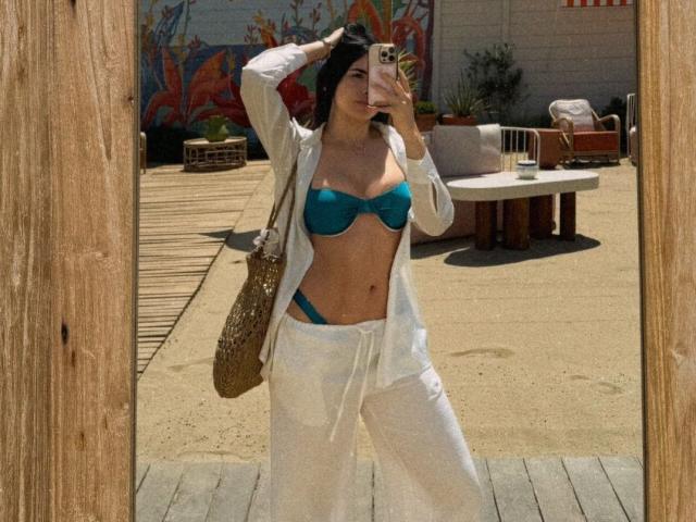LolaHeaven con reggiseno color turchese e pantaloni bianchi, con il seno scoperto, che tiene un telefono, capelli neri ondulati, pelle abbronzata, camicia bianca aperta, specchio con cornice di legno, borsa marrone, patio esterno..