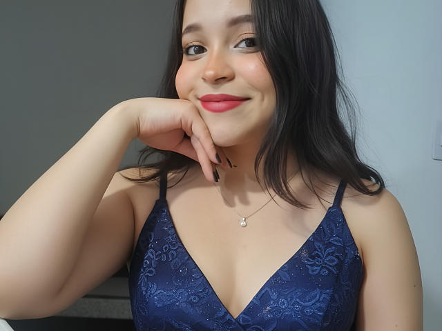 SamyMancini with black hair, red lipstick, blue lace bra, and a cute smile, resting her chin on her hand, showing her tits and a small necklace.