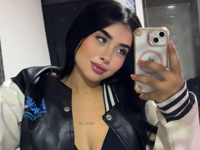 ValerySilva with long black hair, pink lipstick, and a black leather jacket, taking a selfie with a pink phone, showing a tattoo above her tits, looking cute.