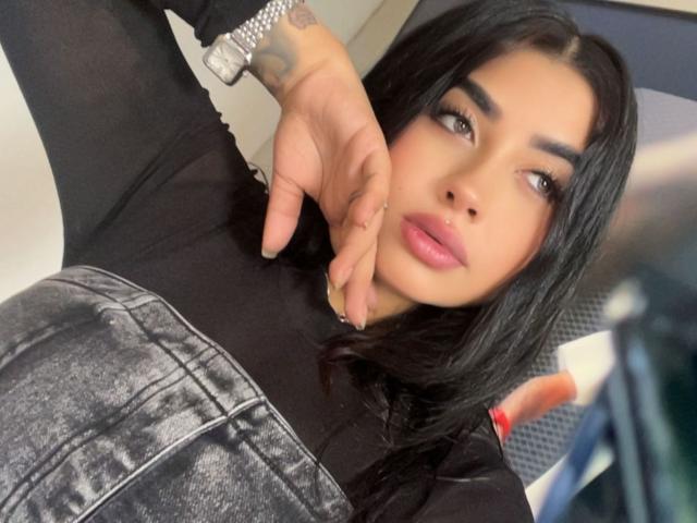 ValerySilva tem cabelo preto, olhos castanhos e lábios rosados. Ela está vestindo uma blusa preta com uma saia texturizada, em tons de cinza e marrom, e seu dedo está perto do rosto. Tem uma tatuagem no pulso..