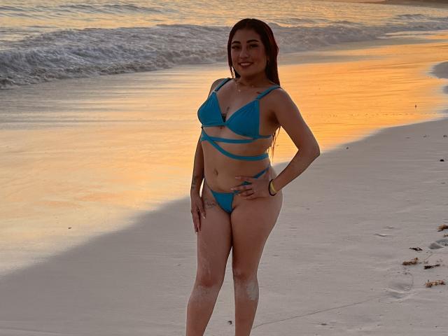 MeganCherryOne se tient sur une plage, vêtue d'un bikini bleu à fines bretelles qui révèle ses seins et ses fesses, avec du sable sur les jambes, et sourit au coucher du soleil..