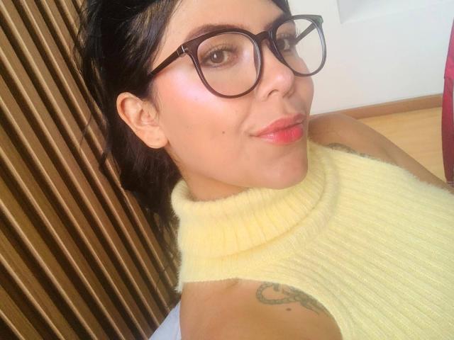 CatalinaReiyna with glasses, red lips, yellow turtleneck, tattoos on her shoulder, and black hair in a ponytail, smiling at the camera. Her ass is partially visible.