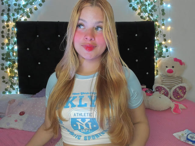ShantalSummer, met lang blond haar en een roze blos op haar wangen, draagt een lichtblauwe en witte Athletic NYC shirt, met een glimlach en roze lippen, zittend op een roze bed..