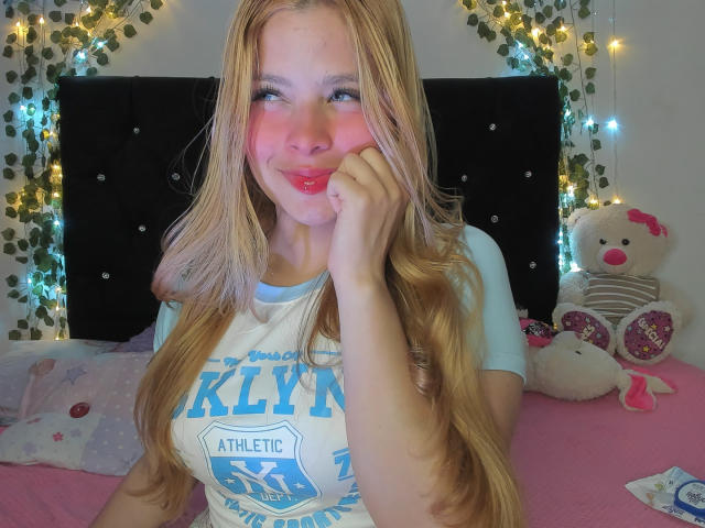 Shantal, een meisje met lang blond haar, een roze blos op haar wangen, blauwe ogen, volle rode lippen, gekleed in een wit en blauw shirt, die haar wang aanraakt en zit op een roze bed met een teddybeer..