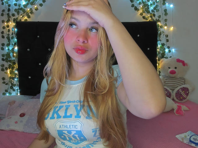Shantal, de cabello rubio, con mejillas rosadas y labios rojos, vestida con una camiseta deportiva azul "Kelvyn", con una mano en la frente, sentada en una cama rosa con un osito de peluche..