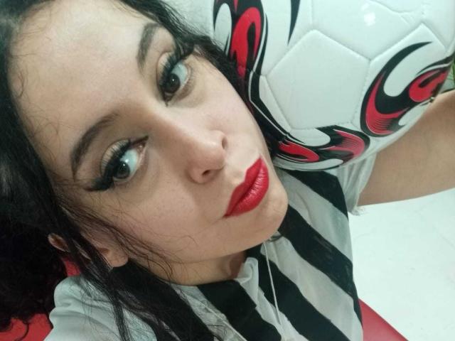 KendraSimons with pouty red lips, black and red striped soccer jersey, holding soccer ball, long black hair, and heavy eyeliner, looking up at camera.