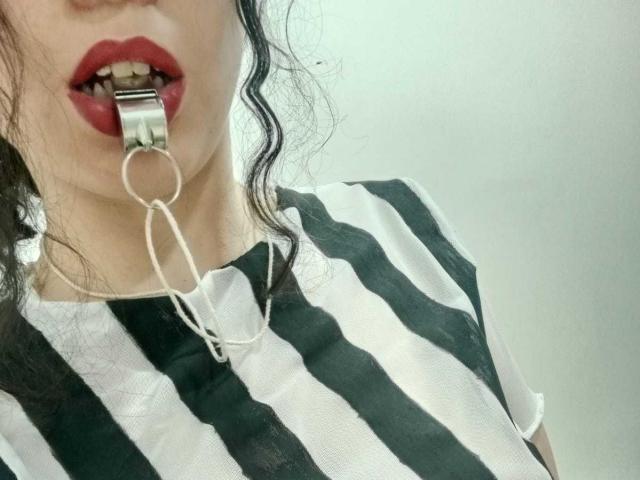 KendraSimons with black and white striped shirt, metal ball gag in mouth, red lipstick, and wavy dark hair, showing upper body and teeth. Her pussy is not visible.