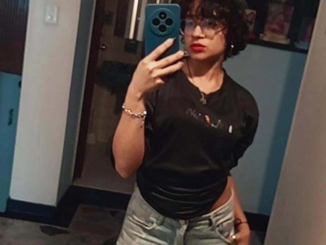 DaryMoon mit lockigem, braunem Haar, Brille, rotem Lippenstift, einem schwarzen T-Shirt und hellblauen Jeans, die ein Selfie mit einem blauen Handy macht. Ihre Hand liegt auf ihrem Hintern..