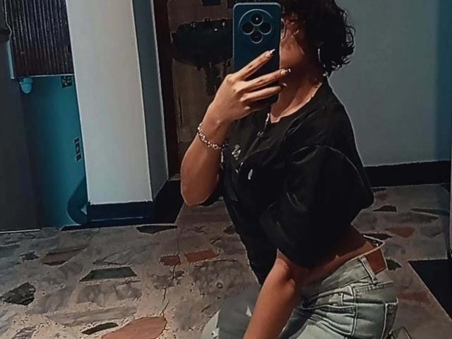 DaryMoon, dark curly hair, black shirt, jeans, kneeling, mirror selfie, holding phone, showing a toned midsection, bracelet, blue phone case, slightly visible butt.