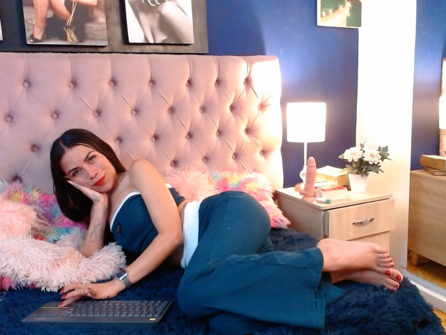 ZoeLux, sdraiata su un letto con una testiera rosa trapuntata, indossa un completo blu e bianco, con il sedere ben in evidenza, piedi nudi, un portatile, un cuscino rosa soffice, una lampada bianca e un sex toy..