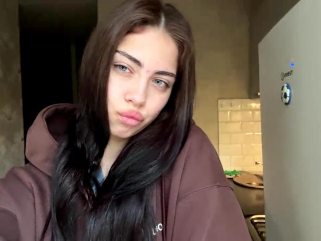 AureaVandale mit langen braunen Haaren, grünen Augen und vollen, rosafarbenen Lippen, trägt eine braune Kapuzenjacke und macht ein Selfie. Im Hintergrund ist eine weiße Tür mit einem Aufkleber eines Fußballballs zu sehen..