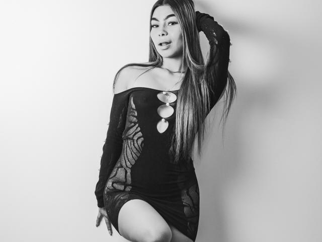 Khaimi, met lang haar, draagt een zwarte jurk met een open schouder en uitgesneden details en ronde knopen. Ze heeft haar hand in haar haar, toont haar borsten en heeft een sensuele glimlach..