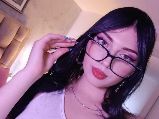 IsabellaaRossi with black hair, glasses, pink eyeshadow, red lipstick, white top, holding nail file, earrings, necklace, light skin, smiling, indoors, slightly blurred.