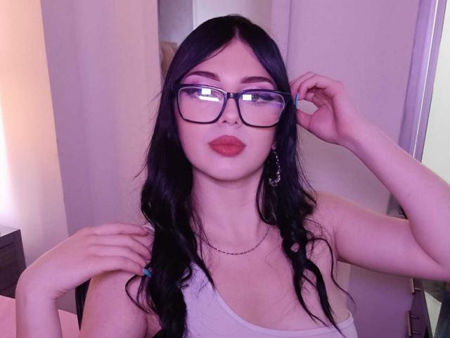 IsabellaaRossi with long black hair, glasses, and red lipstick, wearing a white tank top, touching her hair, showing her tits, and wearing a necklace in a purple-lit photo.