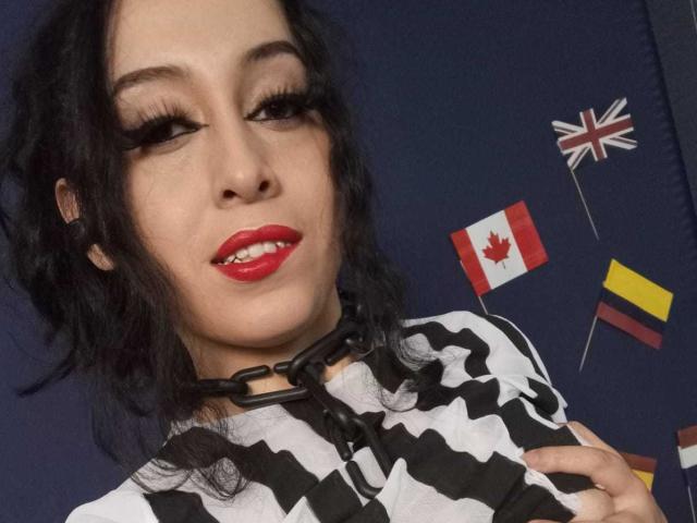 KendraSimons with dark wavy hair, red lipstick, and black/white striped shirt, wearing a black chain choker, smiling with visible teeth, and flag decorations in the background.
